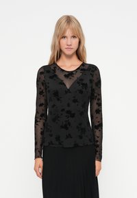 Schwarzes Langarmoberteil aus transparentem Stoff mit floraler Samtstruktur. Mit V-Ausschnitt und figurnaher Silhouette, über einem schwarzen Rock getragen.