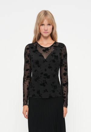 Crni dugosleeve top napravljen od prozirnog materijala s cvjetnim baršunastim teksturom. Ima V-izrez i usku siluetu, složen preko crne suknje.
