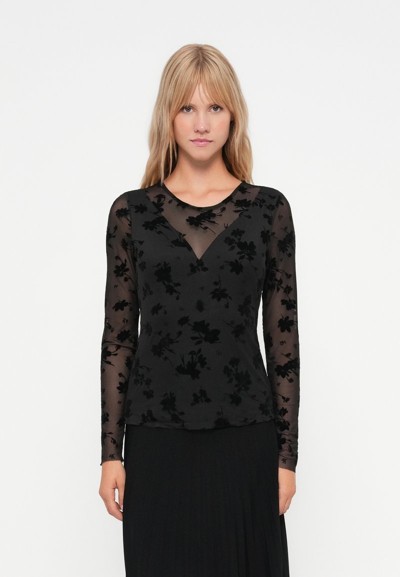 Schwarzes Langarmoberteil aus transparentem Stoff mit floraler Samtstruktur. Mit V-Ausschnitt und figurnaher Silhouette, über einem schwarzen Rock getragen.