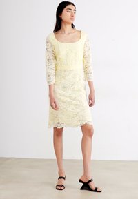 Robe en dentelle jaune pâle avec des manches trois-quarts, un décolleté carré et un ourlet festonné. Associée à des sandales à talons carrés noires.