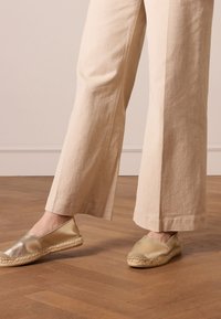 Metalen gouden espadrilles met een gestructureerde bovenkant en een geweven jutesool, gecombineerd met lichtbeige brede broek. Houten vloeren op de achtergrond.