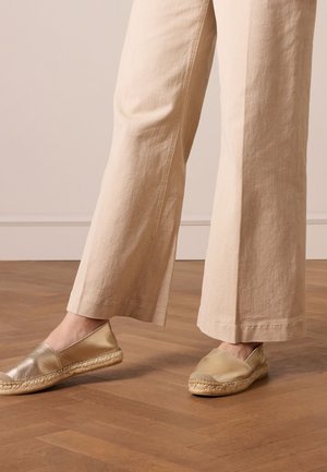 Metallic gold espadrilles with a textured upper and woven jute sole paired with light beige wide-leg pants. Wood flooring background.