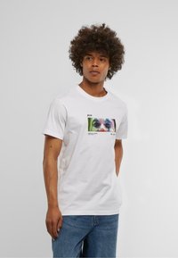 Hvid bomulds T-shirt med et farverigt grafisk print på forsiden. Korte ærmer, rund halsudskæring og afslappet pasform. Blå denim jeans er på.