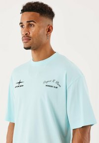 Homme avec des cheveux courts et bouclés, des anneaux au nez et une boucle d'oreille en croix portant un t-shirt bleu clair avec le texte et le logo "Project X Paris Members' Club".