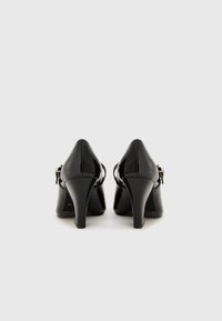 Chaussures à talons hauts en cuir verni noir avec un bout pointu, finition brillante et un petit strap accent près de la cheville. Design à talon.
