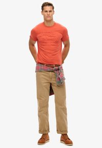 Men's orange embossed graphic t-shirt, khaki pants, plaid shirt tied at the waist, brown leather boots, casual look with neutral background.