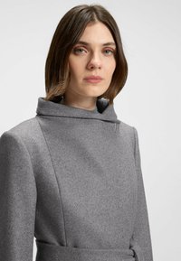 Cappotto grigio in lana con colletto alto, dettagli di cuciture inclinate e un accento laterale a spilla. Texture morbida con una silhouette strutturata.