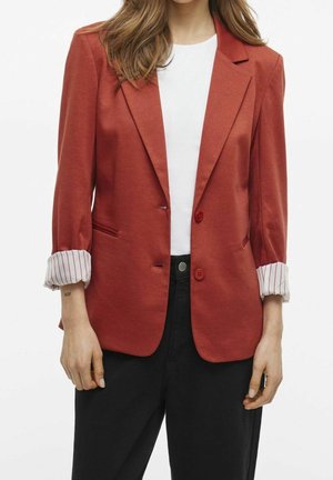 Femme portant un blazer couleur rouille avec des poignets rayés retroussés sur un haut blanc et un pantalon noir, debout devant un fond blanc.