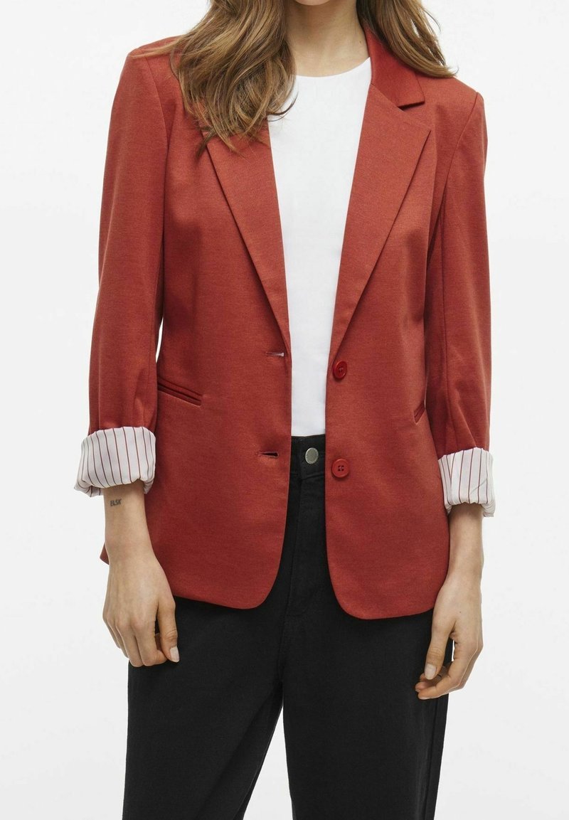 Femme portant un blazer couleur rouille avec des poignets rayés retroussés sur un haut blanc et un pantalon noir, debout devant un fond blanc.