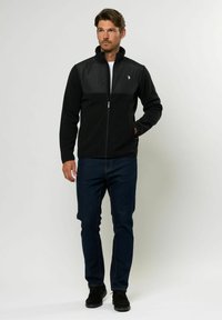 Muž v čiernej vetrovke so zipsom a logom, tmavomodrých džínsoch a čiernych topánkach, stojaci na bielom pozadí.