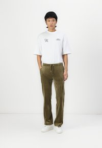 Olivegrüne, weiche Velours-Jogginghosen mit einem Kordelzugbund und seitlichen Paspelierungen, kombiniert mit einem lässigen weißen T-Shirt mit Grafik und weißen Sneakers.