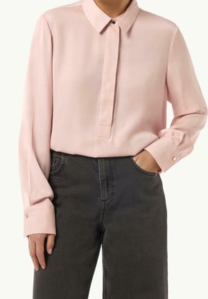 Kvinde iført en lyserød bluse med lange ærmer og skjult knaplukning samt sorte jeans med høj talje, den ene hånd i lommen.