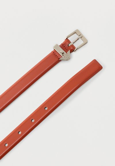 Καφέ δερμάτινη ζώνη με ασημένια αγκράφα και μεταλλική διακοσμητική λεπτομέρεια σε σχήμα αετού κοντά στις τρύπες του κλεισίματος σε απλό φόντο.