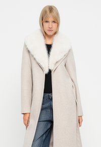 Palton bej deschis cu guler de blană artificială albă, mâneci lungi și un design deschis în față. Purtați peste un top negru și blugi albaștri.
