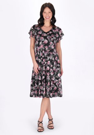 Kobieta stojąca i uśmiechnięta, ubrana w czarną, kwiecistą sukienkę do kolan z falbanowymi rękawami oraz czarne sandały na cienkich paskach.