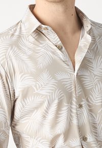 Vaatevaatteet khaki-värisessä lyhythihaisessa paidassa, joka on koristeltu valkoisilla palmulehtikuvioilla. Paidassa on napillinen kaulus ja kaksi näkyvää nappia etuosassa. Kangas on sileä ja teksturoidun tuntuinen.