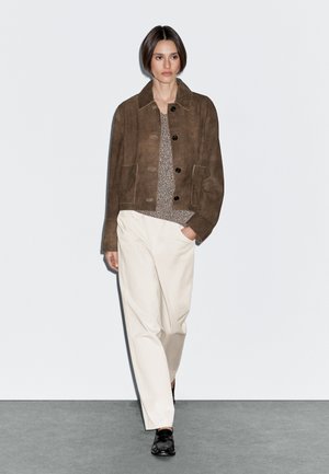 Brown suede jacket with a collar, featuring buttons and front pockets, paired with a textured gray sweater and cream-colored pants. Black shoes.