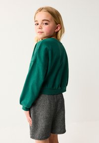 Zelený krátky sveter s širokými rukávmi a tesným lemom, v kombinácii so šedými kraťasmi s houndstooth vzorom, stojaci pred jednoduchým pozadím.