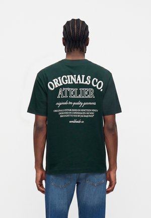 Personne avec des cheveux tressés portant un t-shirt vert foncé avec le texte blanc "Originals Co. Atelier" dans le dos et un jean bleu.