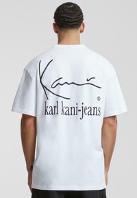 Biela bavlnená tričko s čiernym podpisovým logom "Kani" a nápisom "karl kani-jeans" potlačeným na chrbte. Krátke rukávy a voľný strih.
