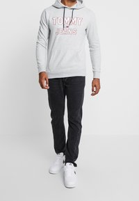 Sudadera gris de algodón con el logo "TOMMY JEANS" en rojo y azul. Combinada con jeans negros y zapatillas blancas con detalles verdes.