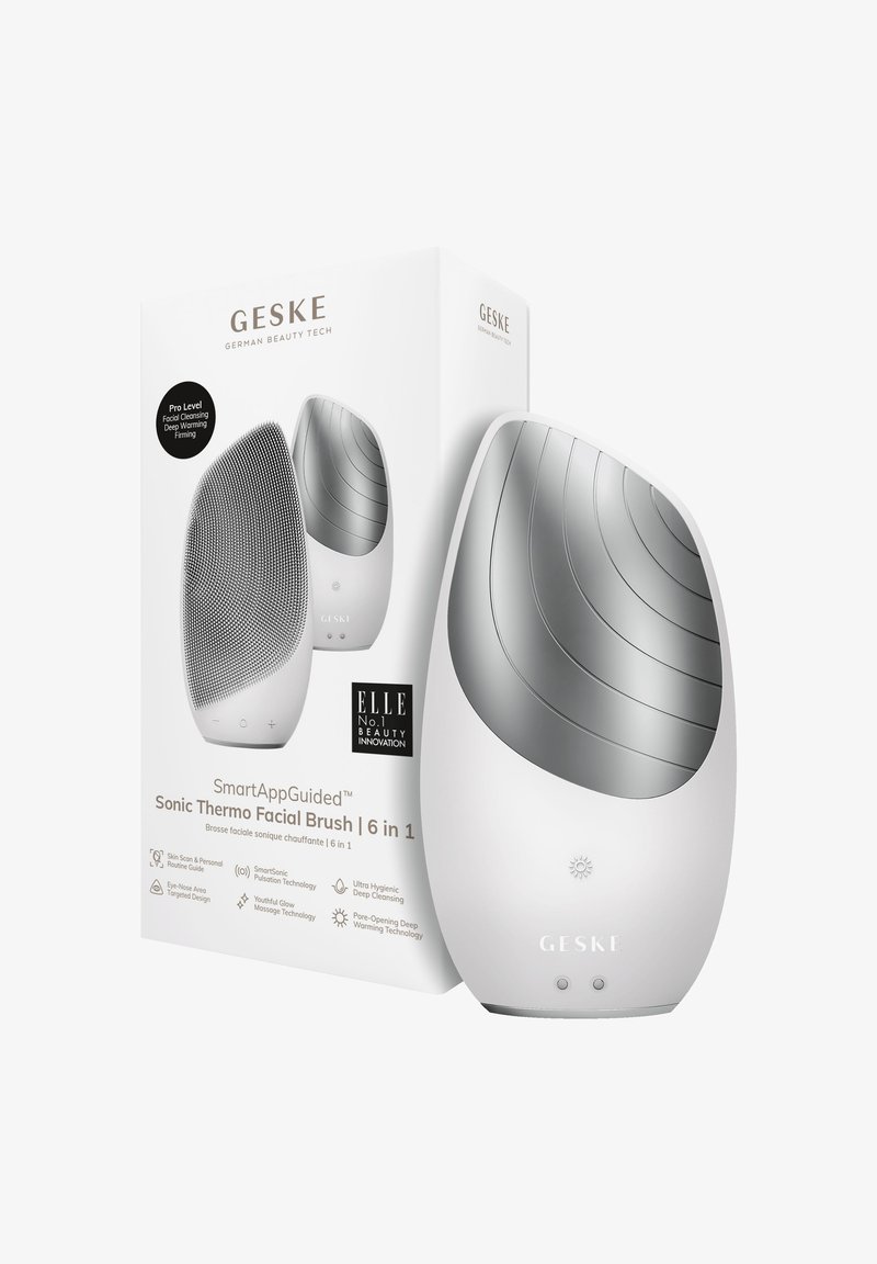 Sonic Thermo Facial Brush i vitt, med en texturerad silikonyta och släta metalliska accenter. Förpackad i en märkeslåda med produktinformation.