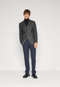 Blazer de tweed gris con un patrón sutil, combinado con un jersey de cuello alto negro, pantalones azul marino y zapatos negros robustos.
