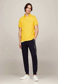 Žltá polo košeľa s krátkymi rukávmi, námornícke jogger nohavice s bielym logom a biele tenisky. Neformálny strih s čistým dizajnom.