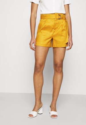 Kvinde iført sennepsgule shorts med bælte, hvid kortærmet bluse og hvide hælede sandaler med åben tå, stående på en grå gulv.
