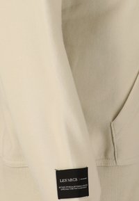 Gros plan sur un tissu beige avec une étiquette de marque rectangulaire noire Les'Arcs cousue près de la couture et du bord de la poche.