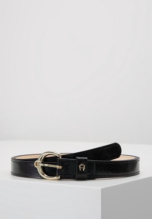 Ceinture en cuir noir brillant avec boucle dorée arrondie et petit logo doré sur la boucle de ceinture, présentée enroulée sur une surface blanche.