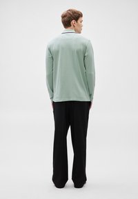 Camisa verde clara de manga comprida com um detalhe subtil na gola, combinada com calças pretas de pernas largas, vista de costas contra um fundo simples.