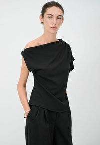 Top nero a spalle scoperte in tessuto morbido, con design drappeggiato e vestibilità ampia. Abbinato a pantaloni neri e un bracciale dorato.