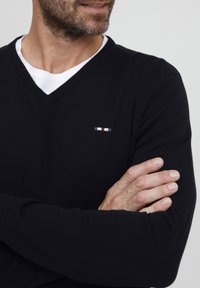 Juodas v-kaklo megztinis, pagamintas iš megztos medžiagos, su įgula rėmeliu. Yra mažas spalvotas logotipas ant krūtinės. Dėvimas ant balto apvalaus kaklo marškinėlių.