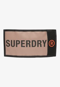 Fascia per testa in tessuto beige con bordi neri che presenta la parola "SUPERDRY" in bold lettere nere e un logo circolare arancione.