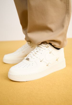 Baskets blanches avec plusieurs petits charms dorés en forme de swoosh Nike, portées avec un pantalon beige sur une surface texturée jaune.