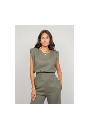 Femme aux longs cheveux bruns portant un haut sans manches vert olive et un pantalon taille haute assorti, accessoirisée de bijoux en or, posant avec la main dans la poche.