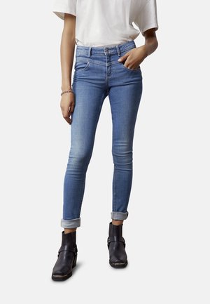 Personne portant un jean skinny bleu clair avec les revers roulés, des bottines noires et une chemise blanche à manches courtes, une main dans la poche.