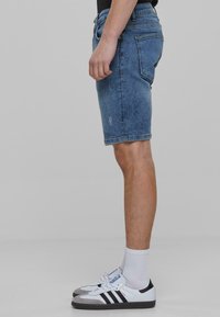 Shorts en denim dans un délavage bleu clair, longueur mi-cuisse avec une coupe droite, comportant des poches et des détails légèrement usés. Associés à des baskets blanches et noires.