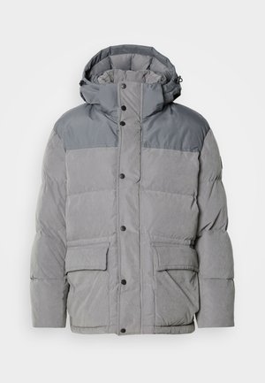 Chaqueta acolchada en gris claro con una sección superior en gris más oscuro, que cuenta con capucha, cierre frontal a presión y dos bolsillos frontales. Textura suave.