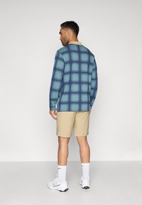 Camisa de manga larga en azul con un patrón de cuadros, cuello beige, combinada con pantalones cortos beige y zapatillas blancas. Tela suave, corte relajado.