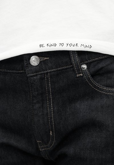 Λευκό μπλουζάκι με την επιγραφή "ΝΑ ΕΙΣΑΙ ΕΥΓΕΝΙΚΟΣ ΜΕ ΤΟ ΝΟΥ ΣΟΥ" στο κάτω μέρος, συνδυασμένο με σκούρο τζιν παντελόνι με ασημένιο κουμπί και ραφές αντίθεσης.