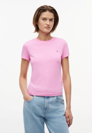 Νεαρή γυναίκα με κοντά μαλλιά φοράει ένα απλό ροζ κοντομάνικο μπλουζάκι και ανοιχτόχρωμα μπλε τζιν, στέκεται με το ένα χέρι στην τσέπη.