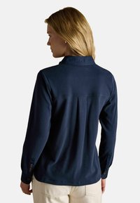 Frau mit blonden Haaren, die ein navy-blaues Langarmshirt und beige Hosen trägt, von hinten vor einem schlichten Hintergrund zu sehen.