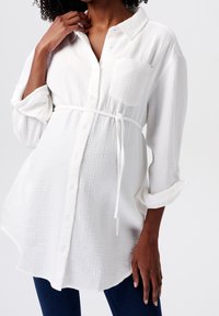 Chemise boutonnée blanche texturée avec une poche poitrine et une taille nouée. Présente des manches retroussées et de légers détails accentués en rose.