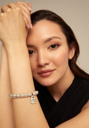 Mujer con cabello largo y castaño, que lleva una pulsera plateada con cuentas y un charm rectangular que dice "UNO de 50", apoyando los brazos cerca de su rostro.