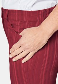 Rood gestreepte broek met elastische tailleband, met een zijzak en een hand die op de stof rust; het materiaal lijkt zacht met een textuurafwerking.