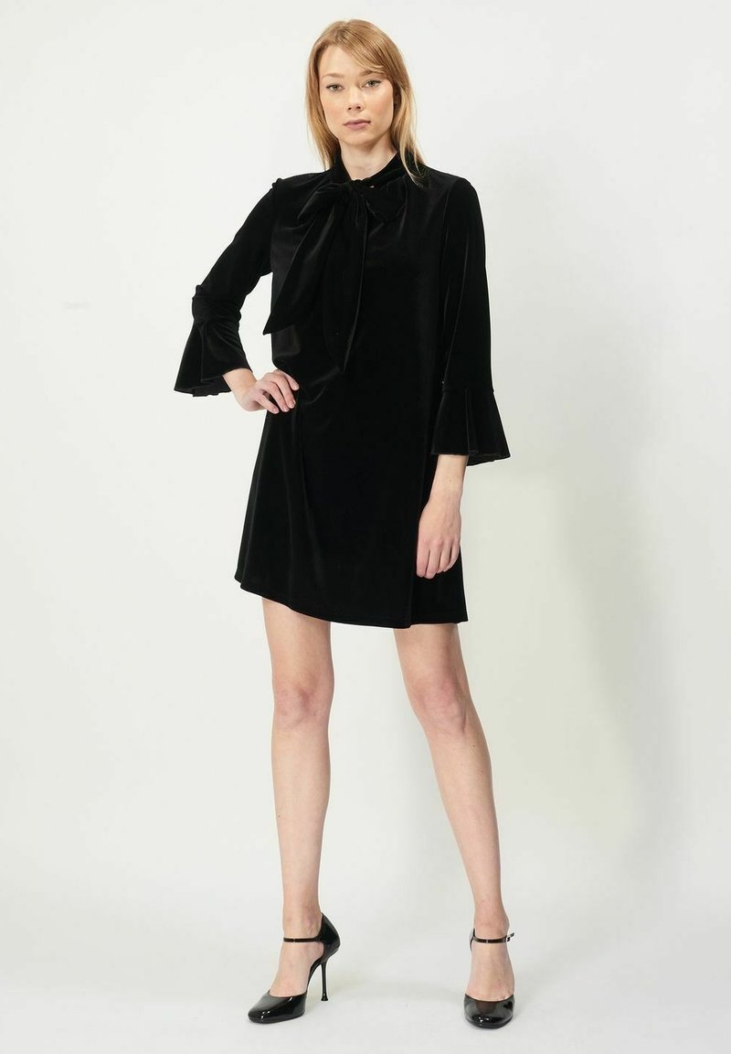 Svart sammetsklänning med knytning vid halsen, långa klockärmar och en utsvängd silhuett, i kombination med svarta högklackade skor.