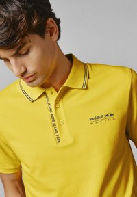 Geltonas polo marškinėlis, turintis vulkaninę apykaklę su tamsiai mėlynais akcentais, dviem mygtukais ir siuvinėtu "Red Bull Racing" logotipu krūtinėje.