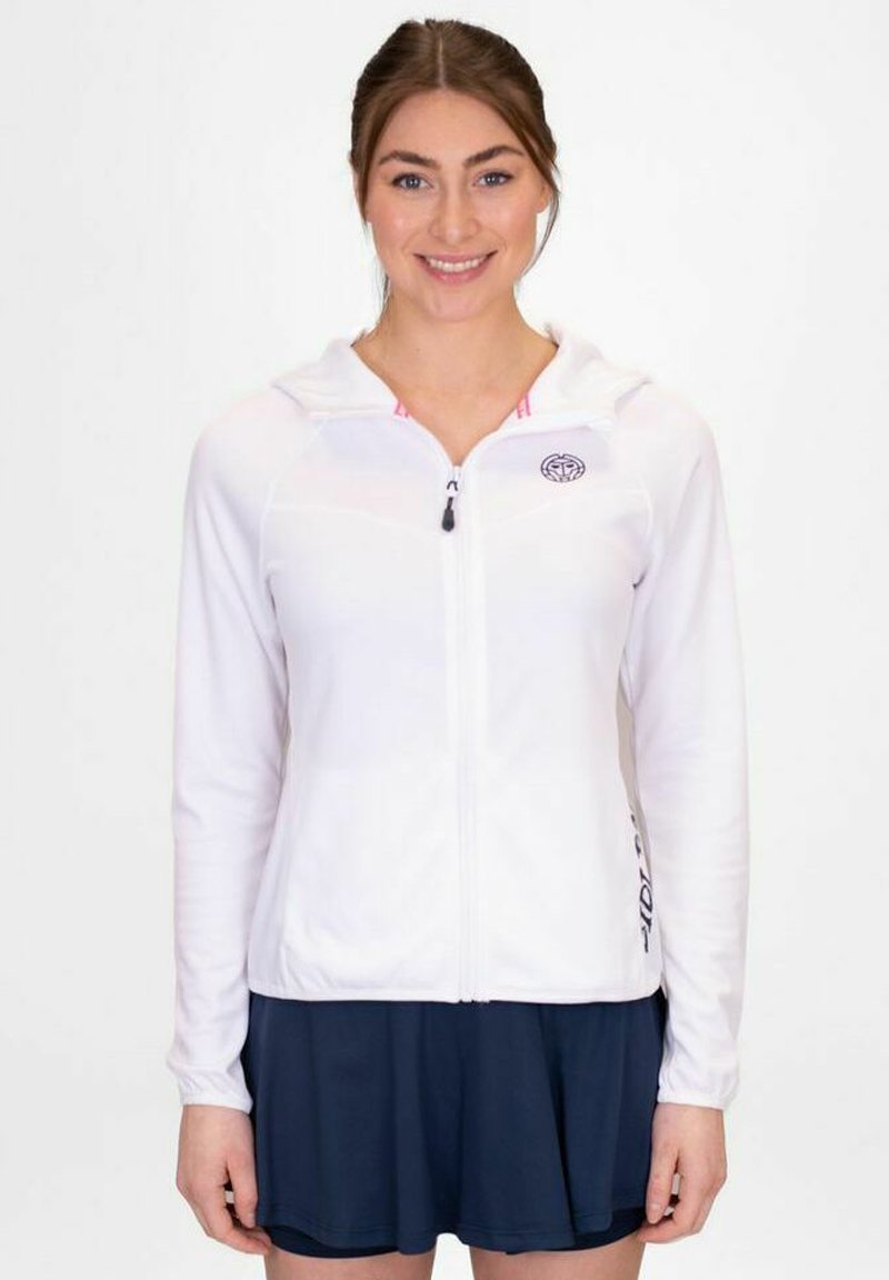 Giacca a zip bianca con cappuccio e maniche lunghe. Presenta un logo sul petto e una fodera interna rosa. Indossata sopra una gonna blu scuro.